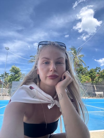 A young woman with blonde hair and light skin takes a selfie outdoors. She is leaning her head on her hand, with a slight smile on her face. She is wearing sunglasses on top of her head and a white top with a cherry print. In the background, there is a blue court, palm trees, and a bright blue sky with fluffy white clouds. The overall impression is one of a casual, sunny day in a tropical location.