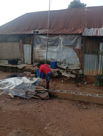 In Karsana 2, Nigeria, a young person in a red t-shirt and denim shorts is seen reaching into a blue plastic container. They are positioned in front of a dilapidated building with a corrugated iron roof and partially covered walls made of mud and concrete. Debris, including wood and plastic sheeting, is scattered around the area. A black basin is visible to the left of the building entrance, and another blue plastic container is near the entrance. The ground is composed of red earth and scattered stones.