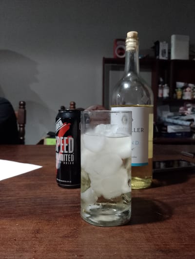 The photograph shows a dark wooden table in Gobernador Julio A. Costa, Argentina. On the table, there is a glass filled with ice and a clear liquid, a black can with the words "SPEED UNLIMITED" in white letters, and a bottle of white wine with a cream-colored label. A white piece of paper is on the left. A mobile phone rests to the side of the bottle. A blurry background shows dark furniture, walls, and other items on the shelves.