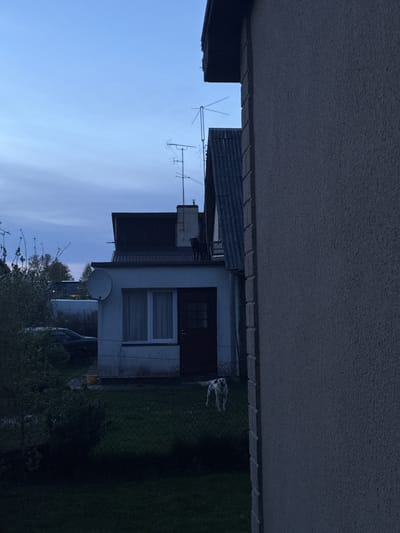 The image depicts a residential area in Ozolnieki, Latvia. A white building with a dark roof is partially visible. A satellite dish and television antennas are affixed to the building. A silver vehicle is parked near the house. A chain-link fence separates the property from an area of green foliage. A portion of an overhanging structure obscures the top of the image. The sky is light blue.