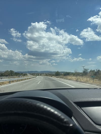 The image depicts a first-person view from inside a vehicle, showing a highway stretching into the distance under a partly cloudy blue sky. The road is paved with asphalt and has white lane markings. A guardrail lines the left side of the road. On the right side, there is dry grass and sparse vegetation, with a green sign labeled "K 589" visible. In the background, hills and some trees can be seen. The car's steering wheel is partially visible in the foreground. The location is identified as the vicinity of Aetokoryfi, Greece.