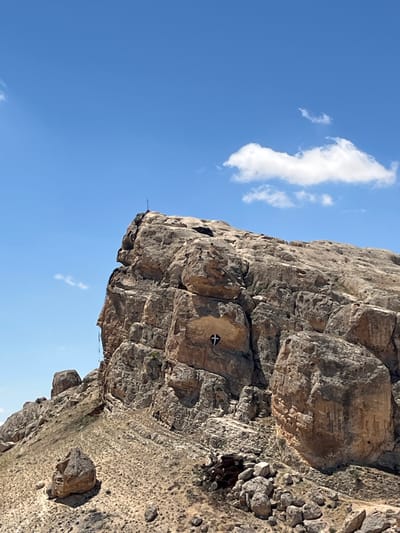 A low-angle, outdoor shot depicts a weathered, light-brown cliff face against a clear blue sky with a small white cloud. The cliff has a rough texture, with various cracks, holes, and rocky protrusions. A small cross is etched or painted onto one of the rock faces. At the top of the cliff are loose rocks. The image is rotated clockwise. The cliff appears to be located in or near Ma'loula, Syria.