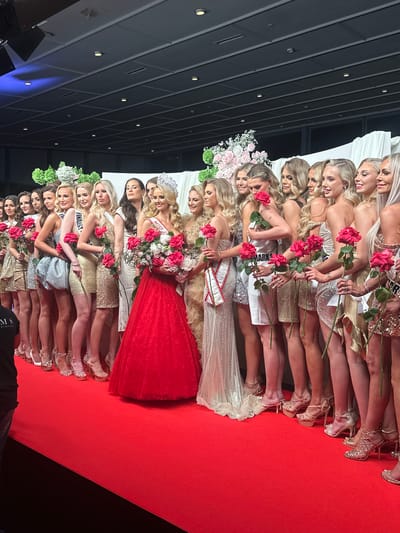 A full-length shot depicts approximately 25 light-skinned women aligned side-by-side on a red carpet, all facing the camera. The women are dressed in various evening gowns and high-heeled shoes. Most hold a single red rose, with the tallest woman in the middle wearing a red dress and holding a bouquet of white and pink flowers. Some are adorned with sashes and crowns.

A dark, modern architectural structure with illuminated panels is visible in the upper left of the frame, providing context to the image. The scene suggests a formal event, possibly a beauty pageant, but it's not possible to determine the location or event details, including whether it is in Skovshoved, Denmark, from the visual information alone.