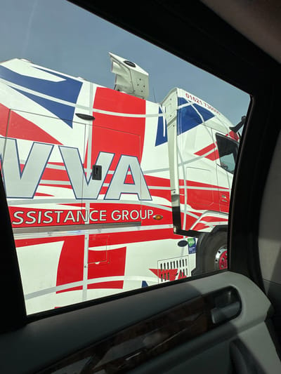 The scene depicts the passenger-side view from within a vehicle, looking outwards at a large articulated lorry (semi-truck) under a clear, light blue sky. The truck, located in Brentwood, United Kingdom, is predominantly white with a distinctive livery featuring red and blue geometric patterns resembling a stylized Union Jack flag. Prominently displayed on the truck's side are the words "VVA ASSISTANCE GROUP" in white capital letters with a blue outline. A phone number, "01621 730000," is visible on the upper part of the truck's cab. A small "HVO" sticker is also present on the lower cab side. The truck features a white, angular air deflector mounted on its roof. Partially visible is a wheel with a red hub. In the foreground, the dark frame of the car window and the vehicle's interior door panel, which includes a gray surface, a simulated dark wood trim accent, and a gray door handle, are present. No individuals are visible in the image.