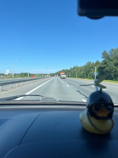 This is a subjective view from inside a vehicle traveling on a highway, likely in the vicinity of Shobashkarkasy, Russia. The view is dominated by a clear blue sky and the road ahead, which has multiple lanes and is bordered by a guardrail and trees. In the foreground, on the dashboard of the car, sits a yellow rubber duck wearing a black helmet and aviator goggles, with a small propeller attached to the helmet. The duck is out of focus. In the distance, several trucks are visible on the highway. The scene suggests a sunny day and travel.