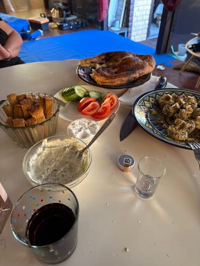 The image depicts a table set for a meal, likely a lunch or dinner, in what appears to be a home setting. The subjects include various food items and dishes arranged on a light-colored tabletop.
Prominently featured is a large, golden-brown pastry, possibly a pie or bread, resting in a dark dish. Next to it, a plate holds sliced tomatoes and cucumbers, suggesting a fresh salad component. A bowl contains a creamy dip with herbs, and a glass container is filled with toasted bread pieces. In the background, a decorative plate holds a portion of food in a creamy sauce, possibly a stew or pasta dish, with a fork reaching into it. There are also two glasses, one empty and clear, and another darker glass containing a beverage, decorated with colorful polka dots. A small silver knife is also visible on the table.
The lighting suggests it is daytime, with sunlight casting subtle shadows. The overall mood appears casual and relaxed, indicating a home-cooked meal or gathering. While specific emotions are not discernible, the arrangement of food suggests an inviting spread.
There is no visible text that can be read clearly in the image. The location context provided is "Столбище, Russia," which helps to situate the scene. The image captures a moment of shared food and dining.