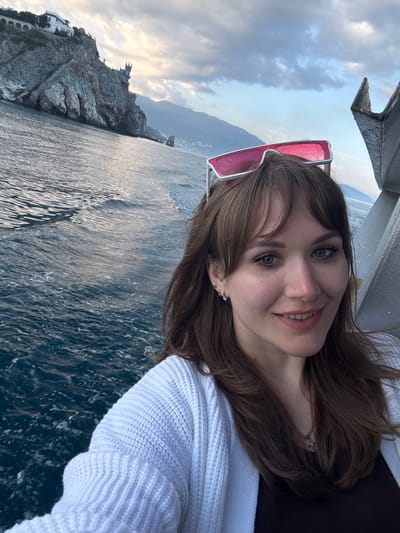 A selfie taken by a fair-skinned woman with brown hair and green eyes, who is smiling slightly at the camera. She is wearing a white cardigan and has pink-framed sunglasses perched on her head. A necklace with a small charm is visible around her neck. The background shows the sea and a rocky coastline with a building atop, suggesting a location in the Гаспринский поселковый совет, Ukraine. The lighting indicates it is daytime, likely early morning or late afternoon.