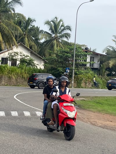 In Walahanduwa, Sri Lanka, two individuals are riding a red scooter on a paved road with white lane markings. The person in the front, wearing a black t-shirt and blue jeans, has a blue helmet. The person in the back, wearing a white and blue striped shirt, has a dark helmet. Both individuals are looking forward. A black SUV is parked on the side of the road behind them. In the background, there are houses with white walls and tiled roofs, surrounded by lush green vegetation and palm trees. A lamppost stands tall to the right of the frame, and a cyclist is visible further down the road. The sky is overcast.
