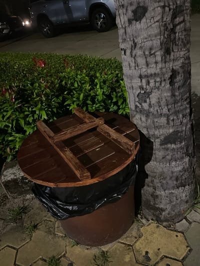 The image captures an outdoor scene at night, likely in a public area given the presence of a trash receptacle. The trash can itself is a large, dark brown cylindrical container, with a black trash bag liner visible at the rim. It is topped with a circular, dark wooden lid, featuring a cross-like wooden reinforcement structure secured with screws.
Adjacent to the trash can is a large palm tree trunk, its rough, textured bark predominantly grey. Behind the trash can, a dense green hedge with small red flowers provides a backdrop. In the distance, the headlights of a vehicle are visible, illuminating a portion of the road and the rear of a light-colored SUV. The ground in the foreground is paved with hexagonal yellow and grey interlocking stones, interspersed with patches of short green grass. The overall lighting suggests it is nighttime, with artificial light sources casting shadows. The scene appears to be in a well-maintained urban or suburban outdoor space.