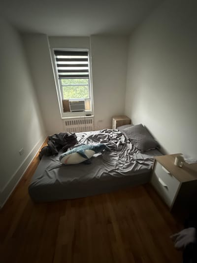 The image depicts a bedroom in the City of Yonkers, United States of America. A mattress, unmade with gray bedding, dominates the center of the room, with a plush shark toy resting on it. To the left of the bed, a pile of dark gray clothing is visible. A window with horizontal blinds is positioned on the far wall, above a white radiator and an air conditioning unit. Adjacent to the window, a small, light brown nightstand is placed. To the right of the bed, a white nightstand with two drawers is situated. On this nightstand, a white mug and some papers are present. The room has light-colored walls and a polished wooden floor. The lighting suggests it is daytime, with natural light filtering through the window, casting shadows across the room.
