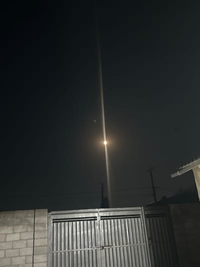 The image captures a nighttime scene in Tokmok City, Kyrgyzstan. The primary subject is the moon, a bright, luminous orb in the dark sky, casting a visible beam of light horizontally across the frame. There are no people visible in the image.
In the foreground, to the right, part of a building is visible. It features a section of grey brickwork at the base, topped by a structure with horizontal grey louvers, likely a window or ventilation grate, with a metal frame. The building's facade to the left is a dark, flat surface. A white pipe or conduit runs diagonally from the top left corner downwards. Thin wires or cables are also visible stretching across the dark sky, creating a sense of urban environment.
The overall atmosphere is calm and serene, dominated by the stark contrast between the bright moon and the dark sky and urban elements. The time of day is clearly night, indicated by the darkness and the presence of the moon. There are no indications of specific weather conditions beyond the darkness. The presence of the building with louvers and the visible wiring suggests an outdoor, possibly residential or commercial, setting in a city. No text is visible in the image.