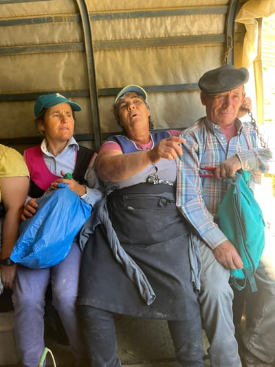 Three individuals, two women and one man, are seated within a semi-enclosed structure, likely a covered transport vehicle or temporary shelter, characterized by a translucent, ribbed plastic sheeting roof and metal framing, under bright lighting. The scene is located in Carrazeda de Ansiães, Portugal.
The woman on the left wears a turquoise baseball cap and a light blue collared shirt with a fuchsia panel. She holds a small green bottle in her right hand and a blue plastic bag rests on her lap. Her gaze is directed towards the right.
The central woman wears a light blue baseball cap with a yellow brim, a dark blue sleeveless top over a checkered shirt, and a dark apron. Her mouth is open, and her right hand is extended, index finger pointing forward.
The man on the right wears a dark flat cap and a plaid shirt (blue, white, and red pattern). His right hand grips a metal chain hanging from above, while his left hand holds a teal backpack. A red-handled tool is visible near his left side. He looks directly forward.
The individuals are seated close together. The floor beneath them appears dark and textured.