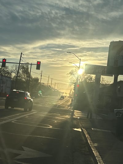 The image depicts a street-level scene at an intersection in Reno, Nevada, specifically near the 7-Eleven and Andy's Taco Shop located at 1195 N McCarran Boulevard. The time is either sunrise or sunset, characterized by a low, bright sun positioned right of center, creating significant backlighting, lens flare, and silhouetted elements. The sky is partially overcast with thin, streaky clouds.

The multi-lane asphalt road displays clear white lane markings, including a left-turn arrow in the foreground. Multiple traffic lights are visible, two on the left and one on the right, all displaying red. A dark-colored SUV is stopped in the foreground, with its taillights illuminated, positioned in a lane marked for straight or left-turn traffic. Other vehicles are visible further down the road, obscured by the sun's glare and atmospheric haze.

On the right side of the image, an individual is standing on the sidewalk near a crosswalk, silhouetted against the bright sun, appearing to wait. Prominent commercial signs for "7-ELEVEN" and "ANDY'S TACO" are visible on the right, indicating active businesses. Utility poles with numerous overhead lines run along both sides of the street, alongside various commercial and residential structures. Trees with sparse foliage are visible on the left side of the road.