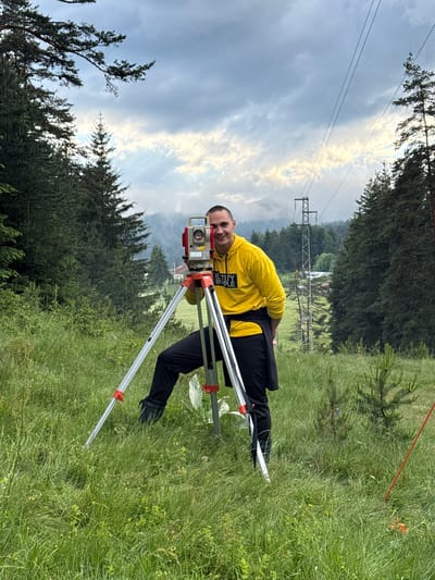 A young man, wearing a yellow hoodie and black pants, stands outdoors in a grassy, treed area, smiling at the camera while operating a surveying instrument mounted on a tripod. The instrument is red and white. The background features a dense forest of evergreen trees, rolling hills, a cloudy sky with patches of sunlight, and power lines supported by utility poles. In the distance, several buildings are visible, suggesting a rural or semi-rural setting, possibly in the vicinity of Yundola, Bulgaria, given the landscape and infrastructure. The overall impression is of a person engaged in outdoor work or a hobby in a natural environment.