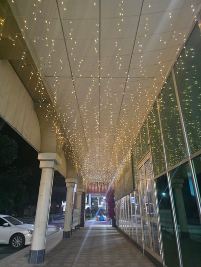 This media file depicts a nighttime scene along an outdoor walkway, likely in Khasavyurt, Russia. The walkway is lined with a building featuring a large, beige-tiled wall adorned with numerous strings of warm, white fairy lights. These lights cascade down the wall, creating a festive and inviting atmosphere. Above the wall, a glass-paneled ceiling reflects the lights and the surrounding environment, hinting at an urban or commercial setting.

The walkway itself is paved with textured, light brown tiles. Several large, cylindrical concrete pillars support the overhead structure, some of which have dark blue bases. A white car is parked to the right of the walkway, its front bumper near one of the pillars. In the background, further down the walkway, there are glimpses of storefronts and signage, with more fairy lights visible in the distance, suggesting a shopping area or entertainment district. Some of the signs in the background are partially legible. One sign, in Cyrillic script, appears to read "СУМКИ ЗАНАВЕСКИ" (SUMKI ZANAVESKI), which translates to "Bags Curtains." Another sign has the word "BUZZ" visible in English.

The time of day is clearly night, indicated by the artificial lighting and the darkness of the surroundings. The weather appears to be clear, as there are no visible signs of rain or snow. The overall mood is one of subdued festivity and urban ambiance, with the soft glow of the fairy lights adding a touch of warmth and magic to the scene.