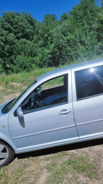 The image shows a silver four-door car parked on a dirt and grass area. The car's right side is visible, with the front and rear passenger doors and windows in focus. The windows are tinted, reflecting the surrounding trees and blue sky. A line of dense green trees forms the background. The car appears to be in a rural or natural setting, possibly near Montagut i Oix, Spain, given the lush vegetation. There are no people visible in the image. The car is angled, suggesting it is parked on a slight incline or on the side of a road. The overall scene is bright and sunny, indicating daytime.
