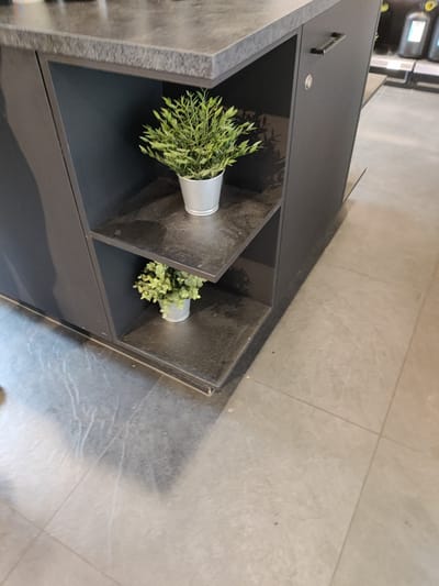 The image shows an interior view of a retail space with tiled flooring. On the left, a dark gray cabinet with open shelves is visible. The top shelf holds a small, potted green plant in a silver-colored pot. The lower shelf also contains a similar small, potted green plant in a silver-colored pot. The cabinet has a dark countertop with a stone-like texture. On the right side of the cabinet, a dark gray door is visible with a black handle and a silver circular lock. In the background, shelves with products are faintly visible, suggesting a commercial setting. The flooring is made of large, light gray tiles. The scene appears to be clean and well-maintained. This image was captured in Eijsden, Netherlands.