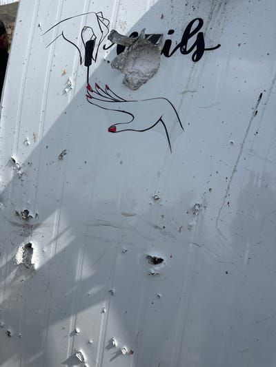 A white corrugated metal or panel surface, located in Dura, Palestinian Territory, is extensively damaged with numerous impact points. The upper section features a graphic of two stylized hands: one hand holds a red nail polish brush, appearing to apply polish to the nails of the second hand. Both hands have red-painted nails. The partial word "ils" from "Nails" is visible in black cursive script in the upper right. The surface exhibits a multitude of small indentations, perforations, and larger pockmarks where the white outer layer is chipped away, revealing a white, crumbly interior material. One significant impact near the word "ils" has torn and warped the surface, leaving a jagged, upward-curling piece of material. The scene is illuminated by strong daylight, casting distinct shadows from the irregularities in the damaged surface and a series of parallel vertical shadows along the left side. No individuals are present.