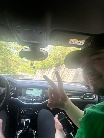 The image depicts the interior of a car. A person wearing a black cap and another person with a green shirt are visible. One of them is holding a smartphone. The car's dashboard features a navigation screen displaying a map. A pair of decorative items are hanging from the rearview mirror. The location is identified as Bilka, Bulgaria.