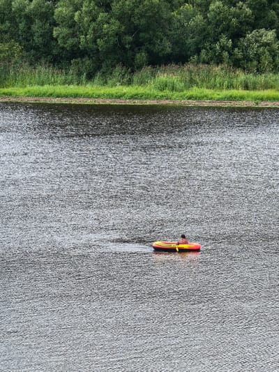 A person is in a yellow and red inflatable boat on a dark, rippling body of water. The person is wearing a reddish-brown shirt and is holding a yellow oar. The boat has the words "EXPLORER 300" written on its side. In the background, a lush green treeline runs along the bank of the water. The location is identified as Staraya Ladoga, Russia.