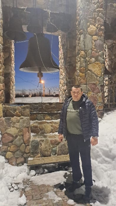 An outdoor scene in the city of Avdot’ino, Russia, depicts a single male subject standing in a snow-covered environment, likely at dusk or night. The man, positioned slightly to the right of the frame, wears a dark quilted jacket over an olive-green long-sleeved shirt and dark trousers. He faces the camera directly. Behind him is a stone bell tower or arched structure, constructed from multi-colored, irregularly shaped stones. Three large bells are visible suspended within the archway, with the largest bell centrally positioned. The base of the structure features stepped stonework, partially covered in snow, and a wooden bench is integrated into the stone wall to the man's immediate left. String lights are affixed to the stone columns of the structure. Beyond the archway, a snow-covered landscape extends under a deep blue sky. A solitary, illuminated street light is visible in the distance, along with faint power lines and the outlines of distant structures or trees. The ground in the foreground consists of a brick pathway, partially covered in patches of snow.