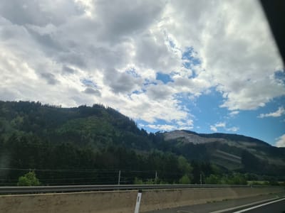 The image shows a mountainous landscape with a highway in the foreground. The mountains are covered in trees, with some areas appearing to have been logged. The sky is cloudy, with patches of blue sky visible. The highway barrier is made of concrete. A white post with a reflective marker is visible on the highway. The scene is likely in Kraubath an der Mur, Austria, as indicated by the context. There are no people or specific actions depicted.