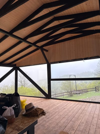 The image presents an interior view in what appears to be a room with a wooden floor, wall, and ceiling. The ceiling features a pattern of wooden planks intersected by dark beams. The room has large windows that offer a view of an outdoor area that seems to be misty, mountainous landscape with a wooden structure resembling a playground. Inside the room, various personal items are visible, including luggage, a hat, a handbag, and a yellow plastic bag, suggesting this may be a guest house or vacation rental. While the prompt requests the location to be identified as Haghartsin, Armenia, there is no visible or readily deducible evidence within the image to verify this claim or link it to a specific location within that city. There are no people visible in the frame, and the items are still.