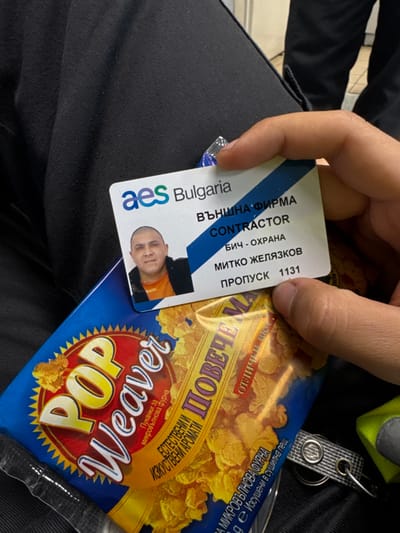 A close-up, first-person perspective image depicts a right hand holding an identification badge and a packet of popcorn. The scene is located within or in the immediate vicinity of the AES Galabovo Thermal Power Plant, near the city of Obruchishte, Bulgaria, as indicated by the "aes Bulgaria" logo on the badge.
The primary subject is a white identification badge with the "aes Bulgaria" logo at the top left. The badge displays "ВЪНШНА ФИРМА" (External Company), "CONTRACTOR," and "БИЧ - ОХРАНА" (Security/Guard). A headshot of a male, identified as "МИТКО ЖЕЛЯЗКОВ" (Mitko Zhelyazkov), is present on the left side of the badge. He has a shaved head and is wearing an orange shirt under a dark jacket. The badge number "ПРОПУСК 1131" (Pass 1131) is printed at the bottom right.
Beneath the badge, partially obscured by the hand, is a blue and yellow packet of "POP Weaver" brand microwave popcorn. Bulgarian text on the packet reads "Пуканки за микровълнова фурна" (Popcorn for microwave oven) and "ЕСТЕСТВЕНИ И ИЗКУСТВЕНИ АРОМАТИ" (Natural and Artificial Flavors), along with "ПОВЕЧЕ" (More) and "отличен вкус" (excellent taste).
The items are held above black fabric, likely pants or a uniform. In the lower right corner, a neon green/yellow strap with a silver keyring is visible. The background is indistinct, showing additional black fabric and a light-colored floor, suggesting an indoor setting.