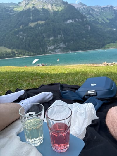 The image shows a scenic view from a grassy bank overlooking a clear blue lake. In the foreground, a person's legs wearing white socks are visible, along with a black blanket, a blue backpack with a TSA lock, and two glasses of drinks. One glass contains a pink beverage, and the other a lighter yellow-green one. The background features a vast expanse of water with a few boats and what appears to be a distant shore with lush green trees and mountains. The sky is a clear blue.

The perspective suggests the person is relaxing and enjoying the view. The bright sunlight and clear skies indicate it is daytime, likely a sunny afternoon. The presence of the lake, mountains, and greenery strongly points to an outdoor recreational setting, consistent with the Glarus, Switzerland location context. The mood conveyed is one of peaceful leisure and enjoyment of nature. There is no visible text in the image.
