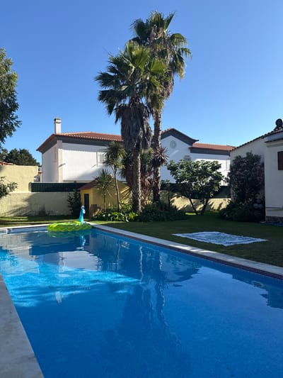 The image depicts a vibrant outdoor scene with a swimming pool as the central focus. The pool is filled with clear blue water, tiled with small, square mosaics, and bordered by a light-colored concrete edge. Floating in the pool is a brightly colored inflatable peacock, adding a playful element to the setting.
Adjacent to the pool is a well-maintained lawn. On the grass, a decorative mat with a mosaic pattern is laid out. In the background, a multi-story building with white walls and a reddish-brown tiled roof is visible. The architecture suggests a residential property, possibly a villa or a house. Lush green trees, including tall palm trees with their fronds swaying gently, and other leafy vegetation surround the building and the pool area, creating a tropical ambiance. The sky is a brilliant, clear blue, indicating a sunny day.
The overall scene suggests a leisurely and relaxing atmosphere, likely during daytime, given the bright sunlight and clear skies. The location cues, such as the architecture and the presence of palm trees, along with the provided context of Seixal, Portugal, contribute to a sense of a warm, Mediterranean or coastal environment. There are no people visible in the image. The time of day appears to be mid-morning or afternoon, as indicated by the strong sunlight casting shadows. The weather is clearly pleasant and sunny.