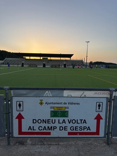 The image depicts a view of a football field at sunset in Vidreres, Spain. In the foreground, a white sign with black text and graphics is prominently displayed. The sign reads "AJUNTAMENT DE VIDRERES" (Town Hall of Vidreres), and below that, "DONEU LA VOLTA AL CAMP DE GESPA" which translates to "Go around the grass field." The sign also indicates "SERVEIS" (Services) with icons for a women's restroom on the left and a men's restroom on the right, each accompanied by a red arrow pointing left and right respectively. The football field itself is green artificial turf, marked with white lines. In the background, a stadium with tiered seating is visible, with the setting sun casting a warm glow across the sky and illuminating the stadium structure. Several individuals are scattered across the field, some appearing to be players, while others are spectating or observing. The overall scene suggests a public sporting facility in use during the evening hours.