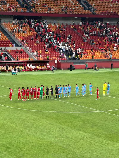 The image depicts a soccer match at the Chongqing Longxing Football Stadium in Longxing, China. The stadium is full of spectators in the stands. Two teams, one in red and the other in blue, are lined up on the field. The red team is lined up with numbers on their backs, including 21, 17, 32, and 33. The two teams are facing each other, likely for the national anthem or pre-game ceremony. Some officials stand between the two teams. There are also people with cameras along the sidelines.