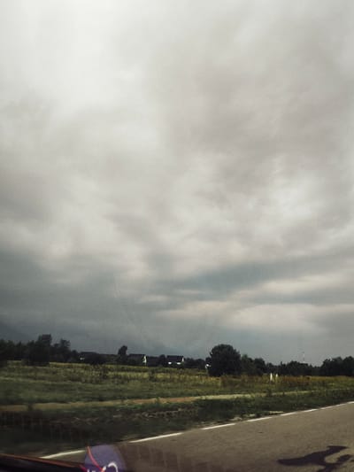 The image depicts a cloudy sky over a rural landscape in Krubin, Poland. In the foreground, a paved road with a white line runs horizontally. The middle ground features a field of green grass and some trees. Several houses with dark roofs are visible in the distance, nestled among the trees. The sky is overcast with dark, heavy clouds, suggesting an impending storm. The overall mood is somber and atmospheric.