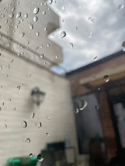 The image captures a close-up view of raindrops on a window. The rain is falling, creating a blurred, overcast sky in the background, suggesting it is likely daytime, possibly with diffused light due to the cloudy weather. The foreground is dominated by numerous water droplets, some small and round, others larger and elongated, distorting the view of what lies beyond the glass.
Through the water-streaked window, indistinct shapes of buildings can be seen, hinting at an urban or semi-urban environment. One of the buildings appears tall and modern, possibly a commercial structure or residential block, given its geometric form and scale. Another part of the scene suggests a more rustic, perhaps older building with wooden beams visible through the rain, adding a layer of contrast to the modern architecture. There's a suggestion of an outdoor space, possibly a balcony or patio, with a blurry object that might be a light fixture or a piece of furniture. The overall atmosphere conveyed by the image is one of a rainy day, creating a somber or introspective mood. There is no visible text in the image. The location context provided is Meljine, Montenegro.