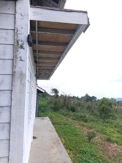 The scene captures the exterior of a building in Man Gyat, Myanmar, under an overcast sky. The left side of the frame features a white, weathered wall constructed from horizontal siding panels, with a prominent vertical corner post exhibiting dark discoloration. A black, multi-finned outdoor light fixture is mounted on this wall. Above, a white roof overhang or eave structure extends, revealing its underside composed of light-toned wooden panels supported by grey metal framing. A narrow, grey concrete path runs along the base of the building. To the right of the path, a dense, natural landscape extends, characterized by a sloping terrain covered in extensive green vegetation, including various grasses, shrubs, and trees, with some areas of drier foliage. Distant hills are visible on the horizon beneath the bright, overcast sky. No individuals or specific activities are present.