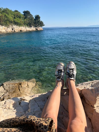 The image depicts a person sitting on a rocky cliff overlooking the Adriatic Sea. The perspective is from above, focusing on the person's legs and shoes, with the sea stretching out to the horizon. The location is Kostrena, Croatia, evident from the rocky coastline and clear turquoise water typical of the region.
The person is wearing white and silver sneakers with black laces. The person has a tattoo on their left ankle. They are wearing a leopard print shirt or jacket. The rocky cliff they are sitting on is composed of light-colored stone with visible cracks and crevices. In the distance, there is a small, tree-covered island. The sea is calm with a visible ripple pattern on the surface. The sky is clear and blue. The scene is illuminated by bright sunlight.