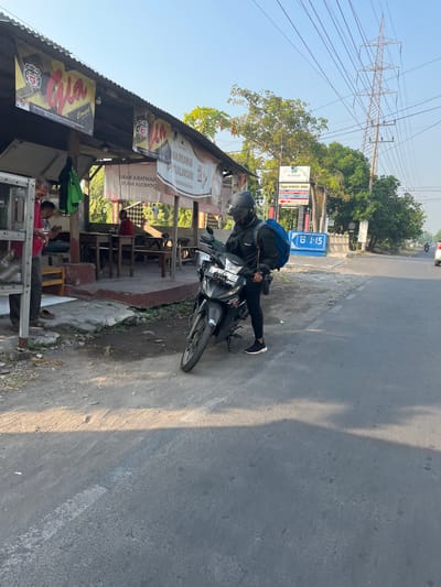 A person wearing a black helmet, black jacket, black leggings, and white sneakers stands next to a black motorcycle. The person is looking down at the motorcycle, which has a blue backpack on the back. The scene is in Sugih Waras, Indonesia, in front of a building with signs that read "Gia" and "Warung Pt. Tulikur." There are wooden tables and benches outside the building. A power line tower is visible in the background. The sky is clear and blue, and the sun is shining.