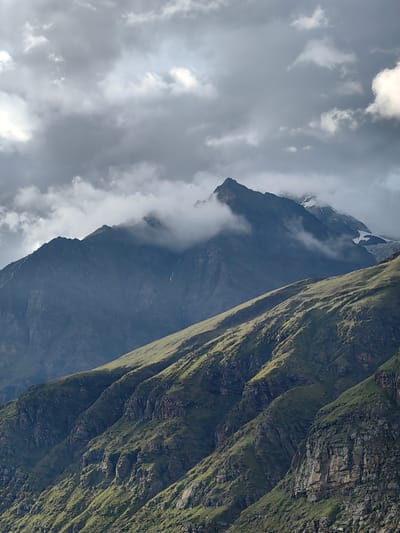 This is a vertical, medium shot photograph of a mountainous landscape, likely taken in Gramphu, India. The foreground features a steep, grassy mountainside with visible rock formations and deep crevices. The slopes are illuminated by sunlight, creating strong contrasts between light and shadow that highlight the terrain's ruggedness. Further back, a darker, more imposing mountain range rises, its peaks partially obscured by dramatic, heavy clouds. Some of these higher peaks show patches of snow and glaciers. The sky is filled with a mixture of dark, turbulent clouds and lighter, brighter areas, suggesting dynamic weather conditions. The overall scene conveys a sense of grandeur and the wild, untamed nature of the mountains.