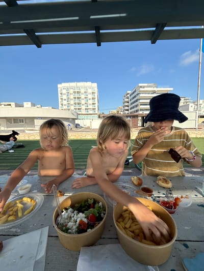 In Nahariya, Israel, three children are gathered around a picnic table, enjoying an outdoor meal. The table is laden with food, including bowls of fries, a salad with feta cheese and tomatoes, and what appears to be bread rolls. One child, wearing a striped t-shirt and a dark bucket hat, is actively eating. Another child, shirtless, is reaching for fries from a large container. A third child, also shirtless, is seated in the foreground, looking down at the table. In the background, a black dog lies on the grass next to a person. Further in the distance, two large, multi-story buildings with balconies are visible against a clear blue sky. The scene is set under a wooden pergola, providing shade.