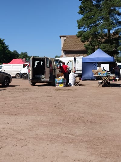 The image captures an outdoor market scene on a sunny day, likely in Irshansk, Ukraine, given the context. The ground is a dry, dirt lot with tire tracks visible. In the mid-ground, a beige van with its rear doors open is parked. A person in a red shirt and black shorts is standing beside the open van, and several cardboard boxes are placed on the ground in front of it. To the right of the van, a large white bag is positioned next to a small wooden stool.
Further to the right, a blue pop-up tent is set up. Beneath the tent, a foldable table displays several items, including boxes of what appear to be bananas. Two people are visible near this table, one standing and the other seated. In the background, more vans are parked, one white with a red top, and another white van with text that includes "АВТО-БР". A building with a tiled roof and stone elements is visible behind the tents and vans. Lush green trees and a clear blue sky complete the environment.