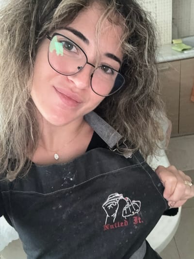 A female individual is depicted in a medium-close-up, frontal portrait within an indoor setting in the city of Kalyvia Thorikou Municipal Unit, Greece. The subject, with light brown and blonde curly hair, is looking directly at the camera with a closed-mouth smile. She wears black-framed, cat-eye style eyeglasses and a delicate gold chain necklace with a small oval pendant. The individual is wearing a dark grey or black apron, visibly marked with light-colored dust or particulate matter. The apron features a white embroidered graphic illustrating two hands, one applying red nail polish to the fingernail of the other, accompanied by the red cursive text "Nailed It." below. The subject's right hand, adorned with a ring on the fourth finger, grasps the lower right section of the apron, near the embroidery. The background reveals a light-tiled wall, wooden cabinetry, and a light green sponge or similar object resting on a counter surface to the upper right.