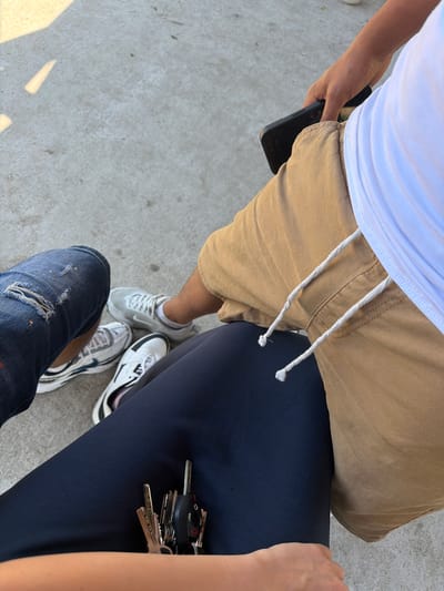 In Dinkata, Bulgaria, a close-up, overhead shot captures the lower bodies and hands of two individuals seated on a concrete surface. One person, on the left, wears distressed denim jeans and white and blue athletic shoes. The other, on the right, is dressed in tan cargo shorts, a white t-shirt, and is holding a black smartphone. Between them, a set of keys is visible resting on a dark blue garment, likely pants or leggings. The background consists of a light gray, textured concrete surface.