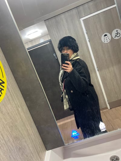A person in a black furry hat and round glasses takes a mirror selfie. They wear a dark, textured coat over a lighter-colored shirt and a patterned scarf. The bathroom setting includes gray walls, a white sink, and signage indicating facilities for disabled people. A "clean" sign is visible on the floor, and a hand soap dispenser is integrated into the mirror.