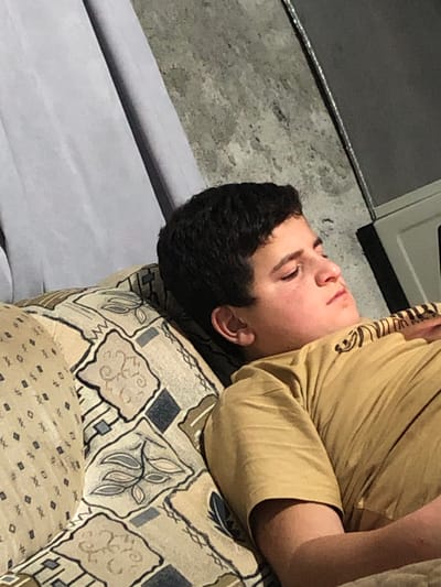 A young male with short, dark hair is depicted lying on his left side on a patterned sofa in an indoor setting in ქვედა სამება, Georgia. He is wearing a tan short-sleeved t-shirt. His eyes are closed, and his head rests on the sofa's armrest or backrest, which is upholstered in a fabric featuring beige, brown, and dark blue geometric and leaf patterns. The background consists of a light grey curtain on the left, a textured grey wall with a mottled appearance in the center-right, and the partial edge of a white-framed, dark-colored rectangular object on the far right.