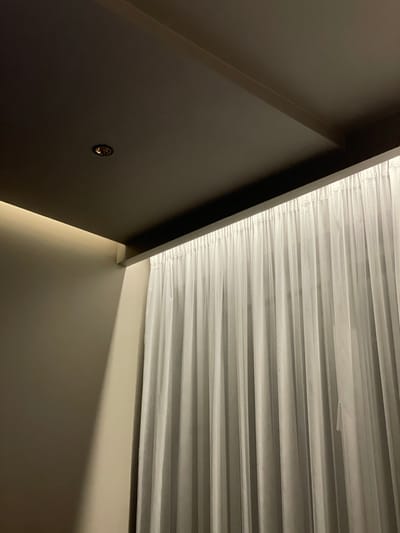 An interior shot showing a portion of a ceiling and wall corner. The ceiling is a light-colored, horizontally pleated material, likely a blind or shade. The walls are painted in a dark, matte color, with a small circular vent visible on one wall. The lighting appears soft and indirect. There is no specific information in the image to definitively locate it in Otopeni, Romania.