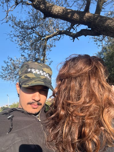 An outdoor, brightly lit daytime image, presumed to be taken in the Menashe Regional Council, Israel, depicts two individuals. On the left, a male with a mustache and medium-dark skin tone wears a camouflage cap with seven white stars on the front and a dark, zipped jacket; he looks directly forward. To his right, the back of a female's head is visible, characterized by voluminous, curly reddish-brown hair. The upper background features a large tree with thick branches and sparse foliage against a clear blue sky, while smaller trees, a light pole, and distant residential-like structures are visible in the left background.