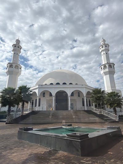 The image shows a modern mosque with white minarets and a prominent dome, set against a cloudy sky. Palm trees are visible in the foreground, suggesting a warm climate. To the right, there is a reflecting pool surrounded by dark stone, and a paved walkway with steps leading up to the mosque entrance. The architecture is contemporary, with clean lines and geometric shapes. The lighting suggests it is daytime, but the clouds obscure direct sunlight, creating a diffused and slightly moody atmosphere. There are no people visible in the frame, and no discernible text. The scene appears serene and contemplative, focusing on the architectural beauty of the mosque and its surroundings. Given the location context of Foz Do Iguaçu, Brazil, the presence of palm trees and the style of the mosque could indicate a significant cultural or religious landmark in the region.