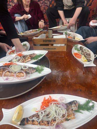 A tilted, medium shot depicts a gathering of people sharing a meal, potentially in Miatli, Russia. The focal point is the grilled fish served on white, fish-shaped platters, garnished with dill, red onion rings, grated carrots, and lemon wedges. Several plates are on a dark wooden table, with a wooden box labeled "by Salmanoff" in Cyrillic script. People are seated around the table, and hands can be seen reaching for the plates. A person on the left is holding a white napkin. The background includes a rug with an oriental pattern. The image suggests a casual dining experience, focusing on the presentation of the food.