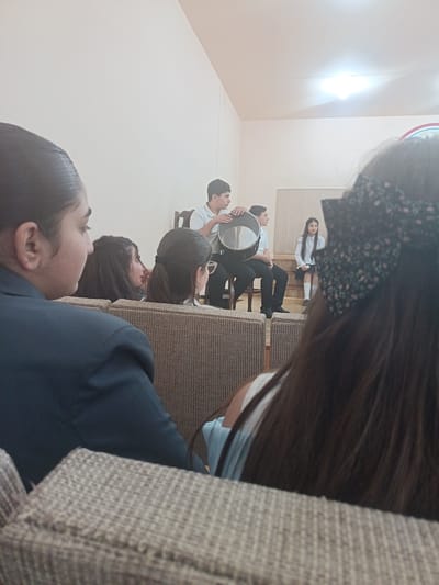 The image depicts a gathering of individuals, likely students, in a room that appears to be a hall or auditorium. In the foreground, several people are seated, viewed from behind. Their hairstyles and clothing suggest a mix of genders and ages. One individual in the far left is wearing a dark jacket. To their right, another person has long, dark hair. Further to the right, a person with glasses and dark hair tied in a ponytail is visible. In the background, a group of young people are standing or sitting, some of whom are dressed in school uniforms. One individual is holding a large drum. Another person, a girl in a white shirt and skirt, is seated on a step or platform. The environment has plain walls, a light fixture on the ceiling, and seating that appears to be upholstered chairs or benches. The overall scene suggests a performance, rehearsal, or an assembly. The city of Nor Hachin, Armenia is mentioned, implying the event might be taking place within an educational institution or community center in that location.