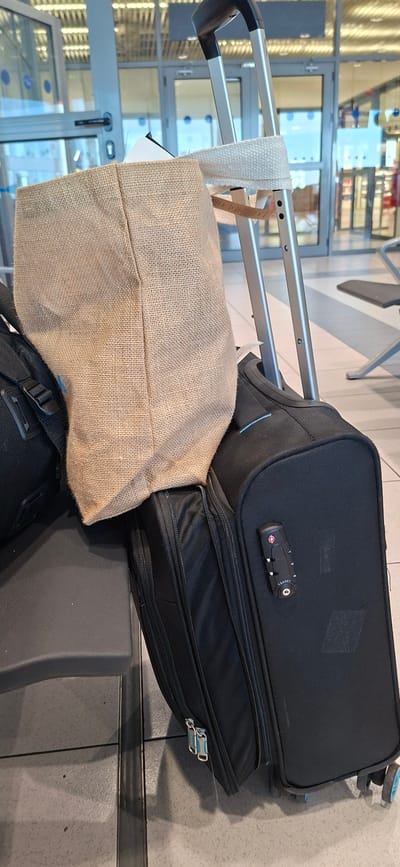 The image shows a close-up view of luggage at an airport terminal. The luggage includes a black rolling suitcase with a TSA-approved lock, a black duffel bag, and a beige burlap tote bag. The burlap tote bag is slung over the handle of the rolling suitcase. In the background, glass doors and a ceiling with fluorescent lighting are visible, indicative of an indoor public space like an airport. The floor is tiled. The scene is set in the city of Rhodes, Greece.